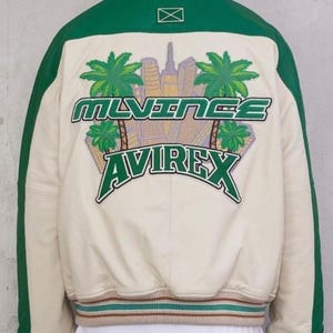 May include: Cream-colored bomber jacket with green accents and a green collar. The back features a graphic with the text "MLVINCE AVIREX" and a cityscape with palm trees. The jacket has green stripes down the sleeves and a striped hem.