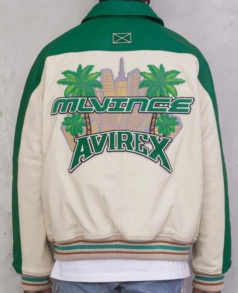 May include: Cream-colored bomber jacket with green accents and a green collar. The back features a graphic with the text "MLVINCE AVIREX" and a cityscape with palm trees. The jacket has green stripes down the sleeves and a striped hem.