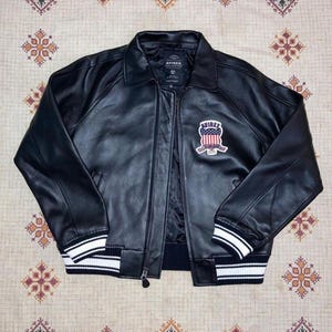 Puede incluir: Chaqueta de cuero negra con cremallera, cuello y pu&ntilde;os y dobladillo acanalados. La chaqueta presenta un parche en el pecho con la palabra "AVIREX" y un dise&ntilde;o con colores rojo, blanco y azul. Los pu&ntilde;os y el dobladillo tienen rayas blancas.