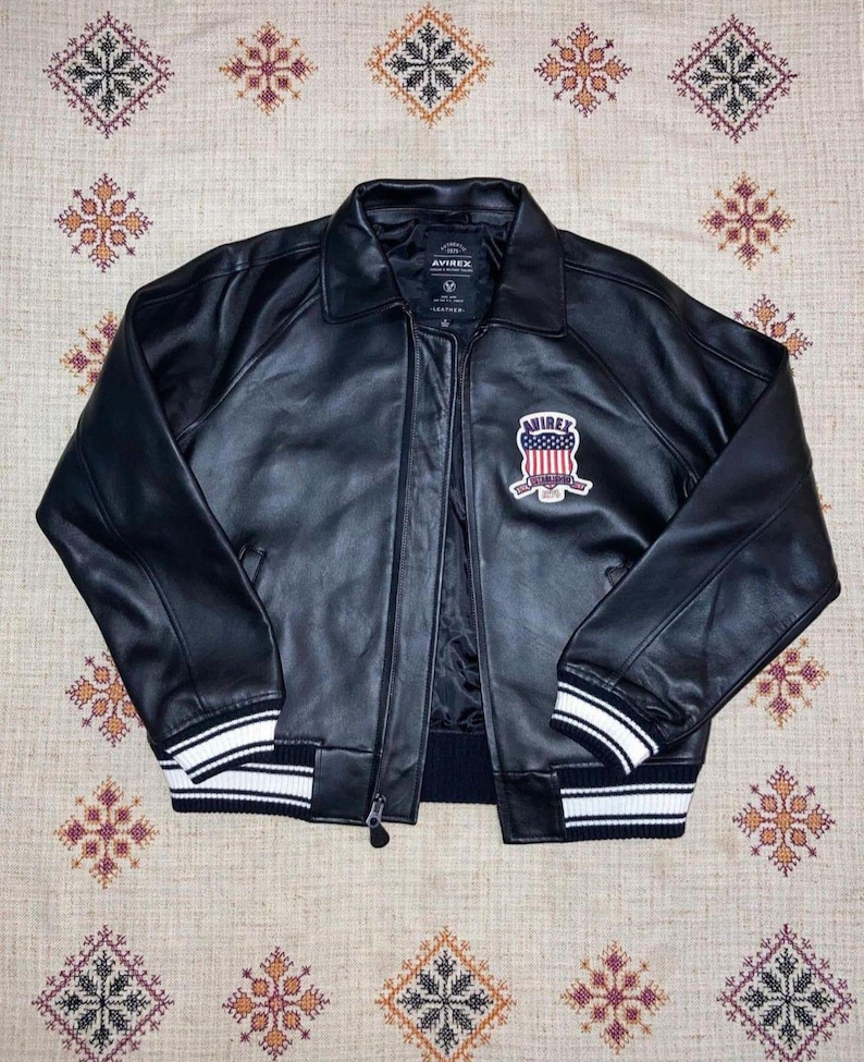 Puede incluir: Chaqueta de cuero negra con cremallera, cuello y pu&ntilde;os y dobladillo acanalados. La chaqueta presenta un parche en el pecho con la palabra "AVIREX" y un dise&ntilde;o con colores rojo, blanco y azul. Los pu&ntilde;os y el dobladillo tienen rayas blancas.