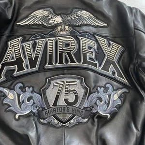 May include: Black leather jacket with embroidered details. Features the word "AVIREX" in large lettering, an eagle emblem, and a shield with the number "75" and the text "AVIATOR'S KING".