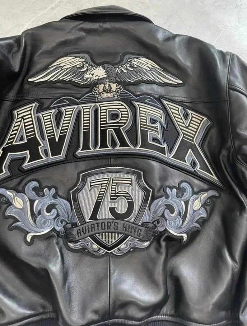 May include: Black leather jacket with embroidered details. Features the word "AVIREX" in large lettering, an eagle emblem, and a shield with the number "75" and the text "AVIATOR'S KING".