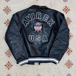 Puede incluir: Chaqueta de cuero negra con la palabra "AVIREX" en letras blancas en la parte superior de la espalda. Debajo, un parche circular con "ALL AMERICANS" y "ESTABLISHED 1975" rodea un gr&aacute;fico de la bandera estadounidense. La palabra "USA" est&aacute; debajo.
