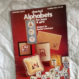 May include: A red booklet titled "charted Alphabets for boys & girls" with designs by Rachel Crissinger. The cover features various cross-stitch projects, including a pillow, a tin can, and a small bag.