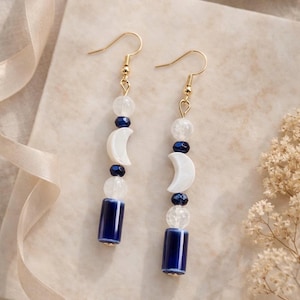 May include: Gold-tone earrings with a celestial theme. Each earring features a gold hook, clear beads, dark blue beads, a white crescent moon, and a dark blue cylindrical bead. The earrings are displayed on a light beige surface.