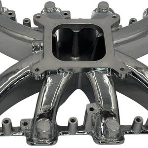 May include: A polished chrome automotive intake manifold featuring a central square carburetor mounting flange and eight curved intake runners that extend outward to connect with engine cylinder heads, showcasing a reflective metallic finish.