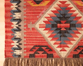 May include: Close-up of a red, geometric patterned rug with a diamond-shaped central design in shades of red, blue, black, and beige. The rug has a fringe along the bottom edge and a textured appearance.