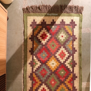 May include: A rectangular jute rug with a geometric pattern in red, green, yellow, and brown. The rug has a green border and a fringed edge. The text "RugVision" is visible on the rug.