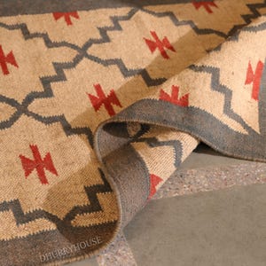 May include: Beige and gray area rug with a geometric pattern. The rug features a repeating design of gray zigzags and red cross-like shapes. The border is a darker gray. The rug is partially rolled up, revealing its texture.