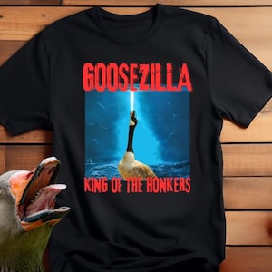 Puede incluir: Camiseta negra con un gráfico de un ganso con un rayo de luz, con el texto "GOOSEZILLA KING OF THE HONKERS" en rojo. Un ganso es visible en la esquina inferior izquierda. La camiseta está sobre un fondo de madera.