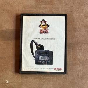 May include: A framed vintage advertisement featuring a monkey wearing a yellow vest and striped pants. Below, a black Aiwa portable cassette player with headphones is displayed. The text reads: IF IT'S NOT AIWA QUALITY, IT'S NOT QUITE RIGHT.