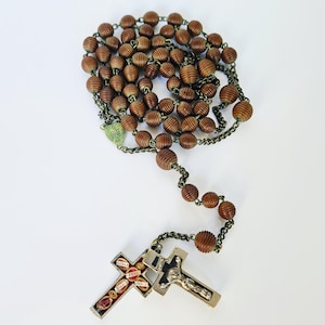 Può includere: Un rosario vintage con perline di legno marrone e una catena di metallo. La croce presenta un crocifisso e intarsi decorativi. È presente anche un piccolo ciondolo verde a forma di cuore.