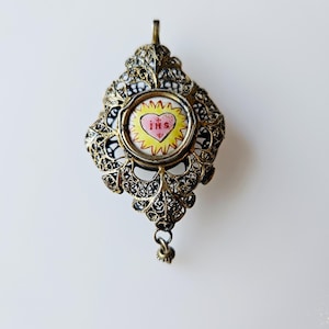Puede incluir: Colgante antiguo adornado con un diseño de filigrana. El colgante presenta una imagen central circular de un corazón con las letras "IHS" en su interior. El corazón es rosa y amarillo, sobre un fondo blanco y negro.