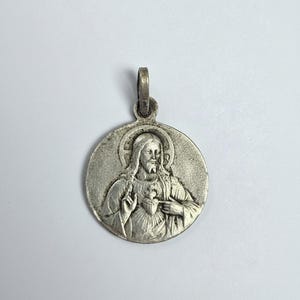 Può includere: Ciondolo argentato raffigurante una figura religiosa con aureola e cuore. Il ciondolo circolare ha un anello per una catena. Il rilievo è dettagliato e il ciondolo ha un aspetto invecchiato.
