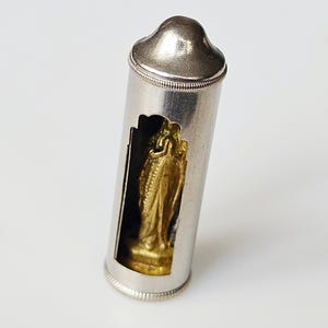 Puede incluir: Un colgante religioso plateado con una estatua dorada visible a través de una abertura rectangular. El colgante tiene una parte superior redondeada y un diseño detallado y texturizado en los bordes.