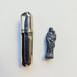 Peut inclure: Un pilulier en métal argenté et une petite statue d'une figure religieuse. Le pilulier est cylindrique avec un dessus arrondi. La statue représente une personne en tenant une autre. Les deux objets sont sur une surface blanche.
