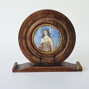 Puede incluir: Un retrato vintage enmarcado en madera de una mujer con un halo, sobre un fondo azul. El retrato es circular, enmarcado en oro, y está dentro de un marco de madera redondo más grande. El marco descansa sobre una base de madera.
