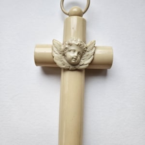 May include: Ivory-colored cross with a cherub design. The cross features a detailed cherub face with wings at the intersection of the vertical and horizontal bars. A loop is at the top for hanging. The cross is made of a smooth, possibly carved material.
