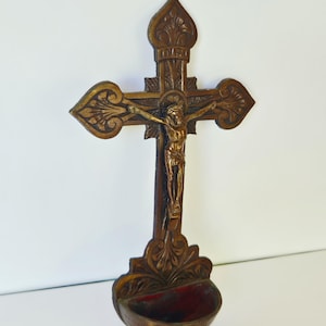 Peut inclure: Une croix en bois sculpté avec une figurine dorée de Jésus. La croix présente des motifs détaillés et un petit élément en forme de bol avec un intérieur rouge à la base. Le design est orné et de thème religieux.