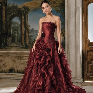 Puede incluir: Un vestido largo sin tirantes en color burdeos intenso con un corpiño ajustado adornado con aplicaciones florales y pedrería, que se transforma en una falda voluminosa compuesta por capas de tela con volantes y textura completa.