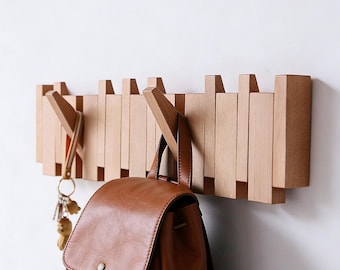 May include: A wooden wall-mounted coat rack with multiple retractable hooks. A brown leather backpack and a set of keys hang from the hooks. The rack is made of light-colored wood and is mounted on a white wall.