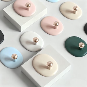 Puede incluir: Una variedad de ganchos de pared ovalados en colores pastel, como rosa, azul y verde. Cada gancho tiene un pomo dorado. Los ganchos están dispuestos sobre plataformas geométricas blancas.