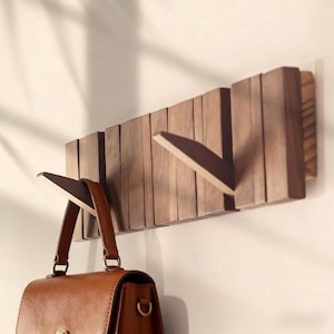 May include: Wooden wall-mounted coat rack with three folding hooks. The rack is made of dark brown wood and has a rectangular shape. A brown leather handbag hangs from one of the hooks.