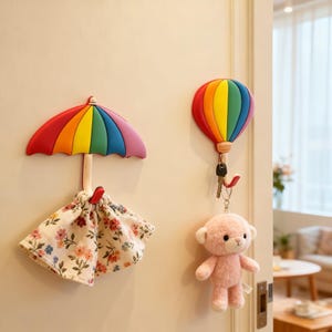 May include: A rainbow-colored umbrella-shaped wall hook with a floral fabric pouch and a hot air balloon-shaped hook. The hot air balloon hook holds keys and a pink teddy bear. Decorative wall hooks.