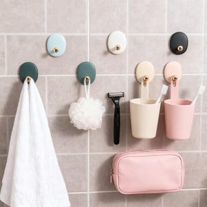 May include: A selection of bathroom accessories against a tiled wall. Includes hooks, toothbrush holders, a razor, a cosmetic bag, and a white towel. The hooks are in various colours, with gold-coloured accents.