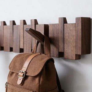 May include: A dark brown wooden coat rack fixed to a wall, with several hooks. The rack has a contemporary design. A brown backpack with a leather strap is suspended from one of the hooks.