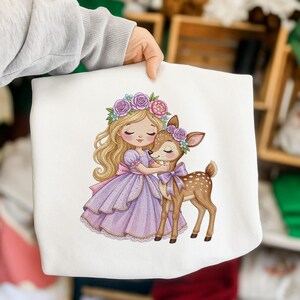 May include: A white sweatshirt featuring a graphic illustration of a blonde character with a floral crown wearing a lavender dress, embracing a spotted fawn that also wears a small purple floral crown and a matching purple bow around its neck.