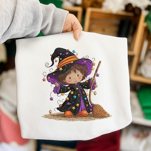 May include: A white sweatshirt featuring a colourful embroidered illustration of a young character wearing a black witch hat and robe decorated with small yellow, blue, and purple stars. The character holds a brown broomstick and wears orange shoes.