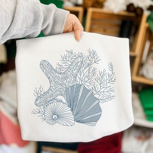 May include: White sweatshirt featuring a blue embroidered design of a starfish, seashells, and coral. The nautical-themed design is centered on the front of the sweatshirt. The sweatshirt is held by a person.
