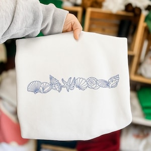 May include: A white sweatshirt featuring a blue embroidered design of various seashells, including a starfish and spiral shells. The design is placed along the bottom edge of the sweatshirt. The sweatshirt is held by a person.