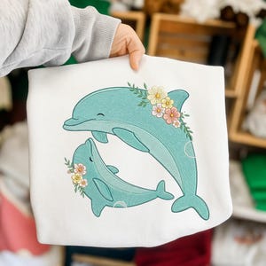 Op de afbeelding: Witte sweatshirt met een geborduurd ontwerp van twee turquoise dolfijnen, elk versierd met bloemenkransen in roze, geel en wit. De dolfijnen glimlachen. De sweatshirt wordt door een persoon vastgehouden.