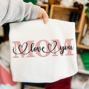 Puede incluir: Sudadera blanca con la palabra "MOM" en letras rosas. Las palabras "I love you" están escritas en cursiva, con corazones reemplazando la letra "o". Un regalo perfecto para el Día de la Madre.