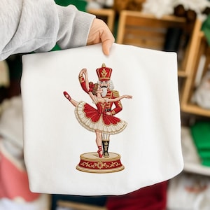 May include: A white sweatshirt featuring an embroidered graphic of a nutcracker soldier in a red uniform dancing with a ballerina in a red and white tutu. The pair stands on a circular red and gold base, captured in a detailed, colourful design.