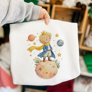 Op de afbeelding: Een wit sweatshirt met een geborduurde illustratie van een jong persoon met blond haar, een gouden kroon, een blauwe jas en een gele sjaal, staand op een kleine planeet omringd door sterren, een maan en een planeet met ringen.