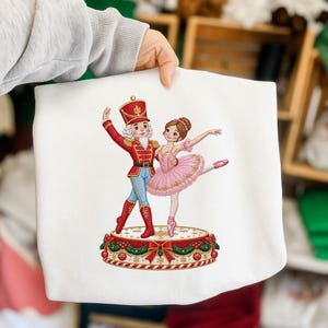 Può includere: Una felpa bianca con un'illustrazione grafica di uno schiaccianoci in uniforme rossa e una ballerina in tutù rosa che danzano su una piattaforma circolare decorata con nastri rossi, ghirlande verdi e motivi a fiocchi di neve dorati.