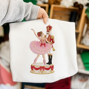 May include: A white sweatshirt featuring an embroidered graphic of a ballerina in a pink tutu and a nutcracker in a red uniform standing on a circular platform decorated with red bows and gold trim. The design is centred on the front of the garment.