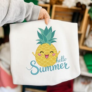 Może przedstawiać: Biała bluza z uśmiechniętym żółtym ananasem, turkusowymi liśćmi i ramionami. Napis "hello Summer" jest wyhaftowany turkusowym pismem. Bluza jest trzymana przez osobę.