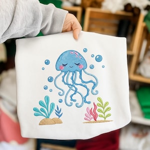 Op de afbeelding: Witte sweatshirt met een geborduurde blauwe kwal met roze accenten, bellen en waterplanten. De kwal heeft een lachend gezicht en lange tentakels, wat zorgt voor een speelse, op de zee geïnspireerde uitstraling.