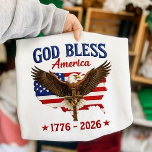 May include: White sweatshirt with the text "GOD BLESS America" in blue and red. An eagle with outstretched wings is centered over a map of the United States, with the American flag design. The sweatshirt also features the text "1776-2026" in red.
