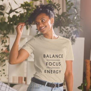 May include: A cream-colored t-shirt with the words "BALANCE FOCUS ENERGY" printed in black. The shirt is a crew neck style and is being worn by a person. The person is smiling and has dark hair.