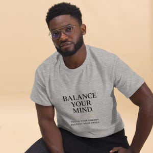 May include: A light gray t-shirt with the text "BALANCE YOUR MIND." below which reads "FOCUS YOUR ENERGY, PROTECT YOUR PEACE." The shirt is worn by a person wearing glasses.