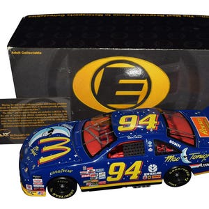 Op de afbeelding: Blauw en geel model van een raceauto met nummer 94, sponsorlogo's en de tekst "Mac Tonight" en "Goodyear". Het model wordt gepresenteerd in een doos met het opschrift "Adult Collectible".