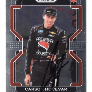 May include: A Panini Prizm trading card featuring a portrait of race car driver Carson Hocevar wearing a black racing suit with Premier Security branding. The card includes a Rookie RC logo, a black autograph, and a dark gray border with red accents.