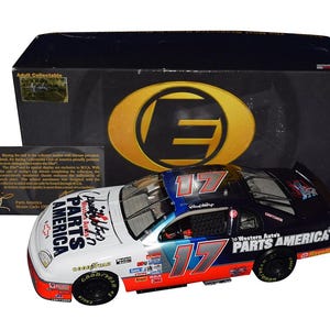 Op de afbeelding: Een gegoten modelraceauto, wit, zwart en rood, met het nummer 17. De auto heeft sponsorlogo's en Goodyear-banden. Op de achtergrond een doos met een groot goudkleurig "E"-logo en een foto van David Welty.