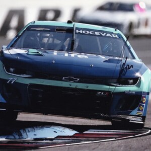 May include: A teal and navy blue race car, number 77, speeds around a track corner. The car has sponsor logos and the name "HOCEVAR" on the windshield. Another car is blurred in the background.