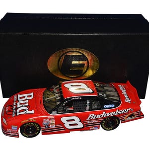 Op de afbeelding: Een rood en zwart model raceauto met nummer 8, Budweiser en Oreo branding. De auto staat voor een zwarte doos met een gouden embleem. Goodyear banden zijn zichtbaar.